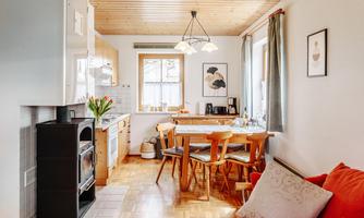A bright kitchen with a dining table and wooden chairs. In the background, there are windows and decorative pictures.