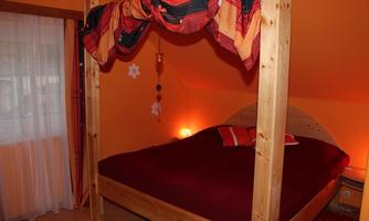 A cozy bedroom with a wooden bed and red bedding. The walls are painted in a warm orange tone, and there are decorative elements like a colorful canopy.