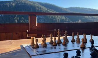 A chessboard with pieces is on a wooden table. In the background, hilly mountains and a blue sky can be seen.