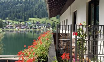 A balcony with blooming flowers and a view of a picturesque lake. In the background, green hills and houses can be seen.