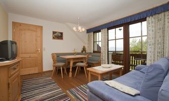 A cozy living area with a blue sofa, a dining table, and wooden furniture. Large windows let in plenty of light and provide a view of nature.