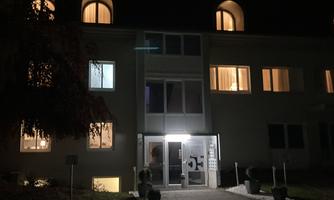 A building at night with illuminated windows. The main entrance is clearly visible and surrounded by green plants.