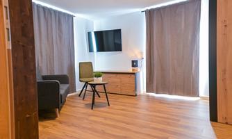 A modern living room with a television on the wall, a cozy armchair, and a table. Large windows with curtains provide bright light and a pleasant atmosphere.