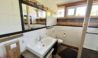 A modern bathroom with white tiles and wooden details. There is a sink, a mirror, and a window that lets in natural light.