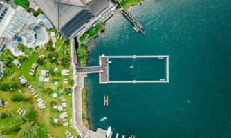 An aerial view of a hotel by the water with a floating pier and a green garden area. In the water, kayaks and swimmers can be seen.