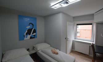 A modern room with two single beds and minimalist decor. A poster of a person hangs on the wall, and there is a window with a view outside.