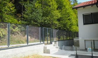 A modern house with a sturdy fence and a green environment. In the foreground, stairs and a small area with wooden planks can be seen.