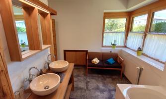 A modern bathroom with two sinks and large windows. There is a cozy seating area and plenty of natural light.