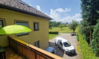 A balcony with a green sunshade overlooking a green meadow. In the foreground, cars are parked and a bright car is parked on the road.