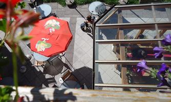 A cozy outdoor area with round tables and a red sun umbrella. In the background, there are plants and a glass building.
