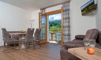 A modern living room with a dining table and chairs. Large windows provide a view of nature and a balcony.