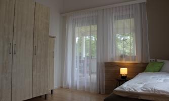 A bright bedroom with a bed, a nightstand, and a wardrobe. Large windows with sheer curtains let in plenty of light.