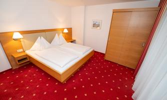 A simple hotel room with two beds and red carpet flooring. The walls are light and there are wooden details as well as a wardrobe door.