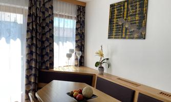 A cozy dining area with a wooden table and colorful fruit. Daylight streams through the windows, and a flower adds a fresh touch to the room.