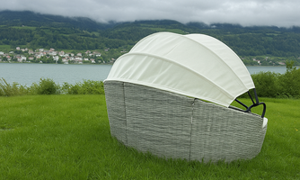 A comfortable lounger with a white sunshade stands on a green meadow. In the background, a lake and wooded hills can be seen.