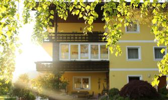 A cozy yellow house with a balcony and large windows. In the foreground, there are green plants and the sun shines through the leaves.