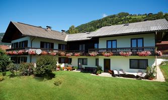 A two-story house with a green garden and balcony flowers. In the background, hills and blue sky can be seen.