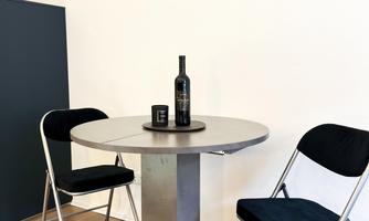 A modern metal table with two chairs stands in a bright room. On the table, there is a bottle and a glass.