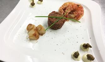 An artfully arranged dish featuring a portion of ground beef, a shrimp, and fried potatoes. The garnish consists of small dollops of capers and fresh chives.