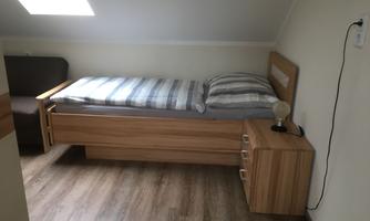 A simple bedroom with a single bed and a wooden dresser.  
There is a cozy armchair and a window for daylight.