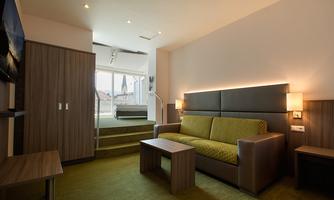 A modern hotel room with a green sofa and a small table. Large windows provide plenty of natural light and a welcoming atmosphere.