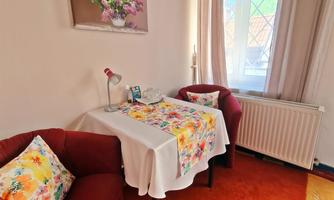A cozy living room with two red armchairs and a small table. On the table lies a floral cloth, and a window lets in a lot of light.