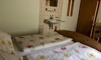 A simple bedroom with two beds and blooming bed covers. On the wall, there is a sink and an open wardrobe.
