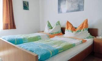 A bright bedroom with a large double bed and colorful bedspreads. A picture hangs on the wall, and the window has orange curtains.
