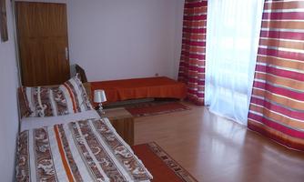 A bright room with two beds and an orange bedspread. Large windows with bright curtains let in plenty of light.