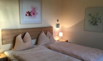 A cozy bedroom with two beds and beige cushions. There is soft lighting and decorative pictures on the wall.