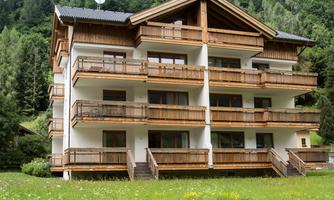 A modern house with wooden balconies in a green environment. Forested mountains can be seen in the background.