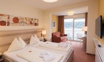 A cozy hotel room with two beds and a television. Through the window, there is a view of the lake and a balcony.