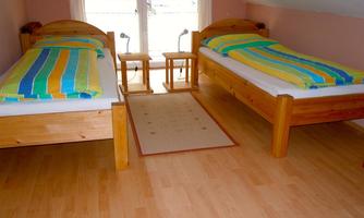 A cozy bedroom with two single beds and colorful bedcovers. The room has wooden flooring and large windows that let in a lot of light.