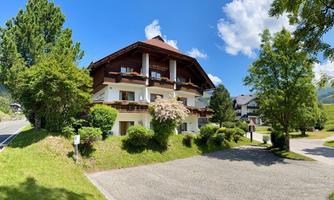 A beautiful house in alpine style with a balcony and a large garden. Surrounded by trees and green grass under a clear blue sky.
