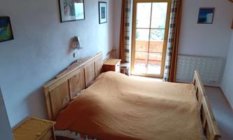 A cozy bedroom with a large double bed and wooden furniture. The balcony door lets in a lot of daylight and offers a view outside.