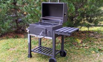 A black grill stands on the lawn, surrounded by trees. The grill has an open lid and a shelf on the side.