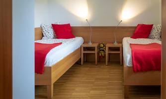 A bright bedroom with two single beds and red pillows. The walls are plain and there is a warm wooden floor.