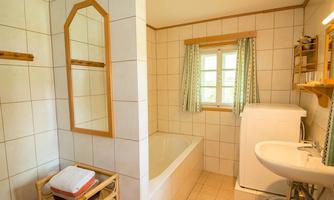 A bright bathroom with a bathtub, sink, and washing machine. The walls are tiled, and there are windows with curtains.