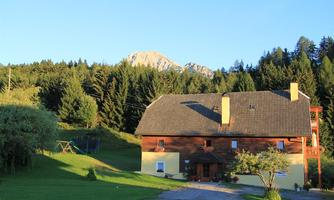 A cozy house amidst trees and green meadow. In the background, majestic mountains can be seen.