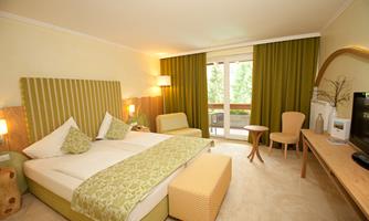 A modern hotel room with a large bed, bright colors, and cozy furniture. Large windows provide plenty of daylight and a view outside.