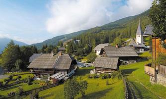 A picturesque mountain landscape with traditional wooden houses and plenty of greenery. In the background, gentle hills and a church tower are visible.