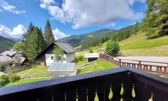 An idyllic landscape with green hills and mountains in the background. In the foreground, typical alpine houses and a balcony with a wooden railing can be seen.
