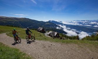 Two cyclists ride along a path in a picturesque mountain landscape. In the background, clouds and a magnificent view of the mountains can be seen.