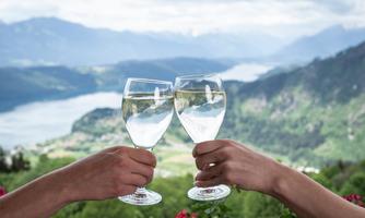 Two glasses of water are clinked together, while in the background a beautiful landscape with mountains and a lake is visible. The scene conveys a sense of joy and relaxation.