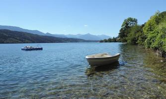 A tranquil lake with clear water and a small boat ramp. In the background are gentle mountains and a bright blue sky.