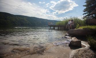 A calm lake with a wooden pier and green shoreline vegetation. Some people are enjoying the view of the water surface and the surrounding hills.