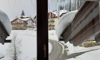 Looking through a window, you see a snowy village with snow-covered roofs. Icicles hang from the eaves and a winding road passes by.