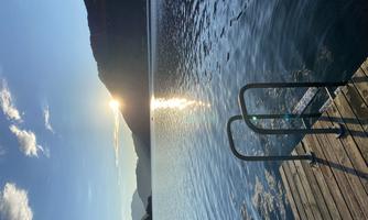 A tranquil lake landscape with gentle mountains in the background. The sun reflects on the water's surface and a dock leads into the water.