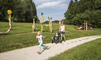 A beautiful park with green meadows and colorful wooden sculptures. A girl happily runs along the path while a woman follows behind with a stroller.