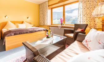 A cozy room with a large bed and a seating area. Large windows offer a view of the mountains and provide plenty of daylight.